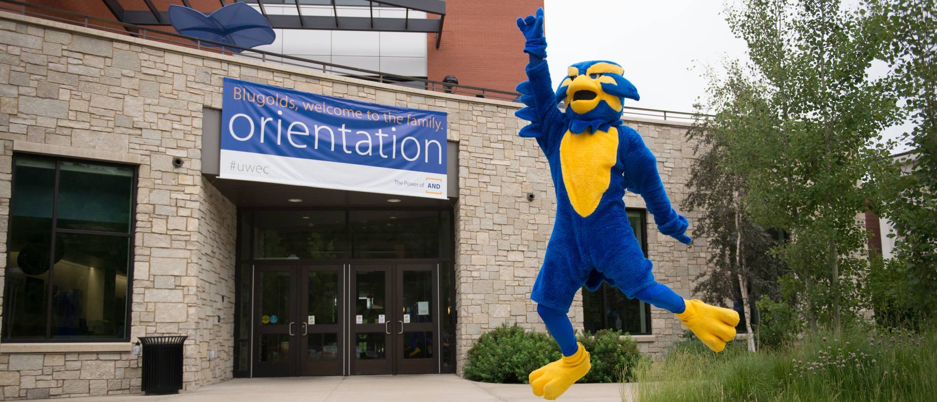 Mascot Blu jumps outside of the Davies Center with an orientation banner hanging from the building.