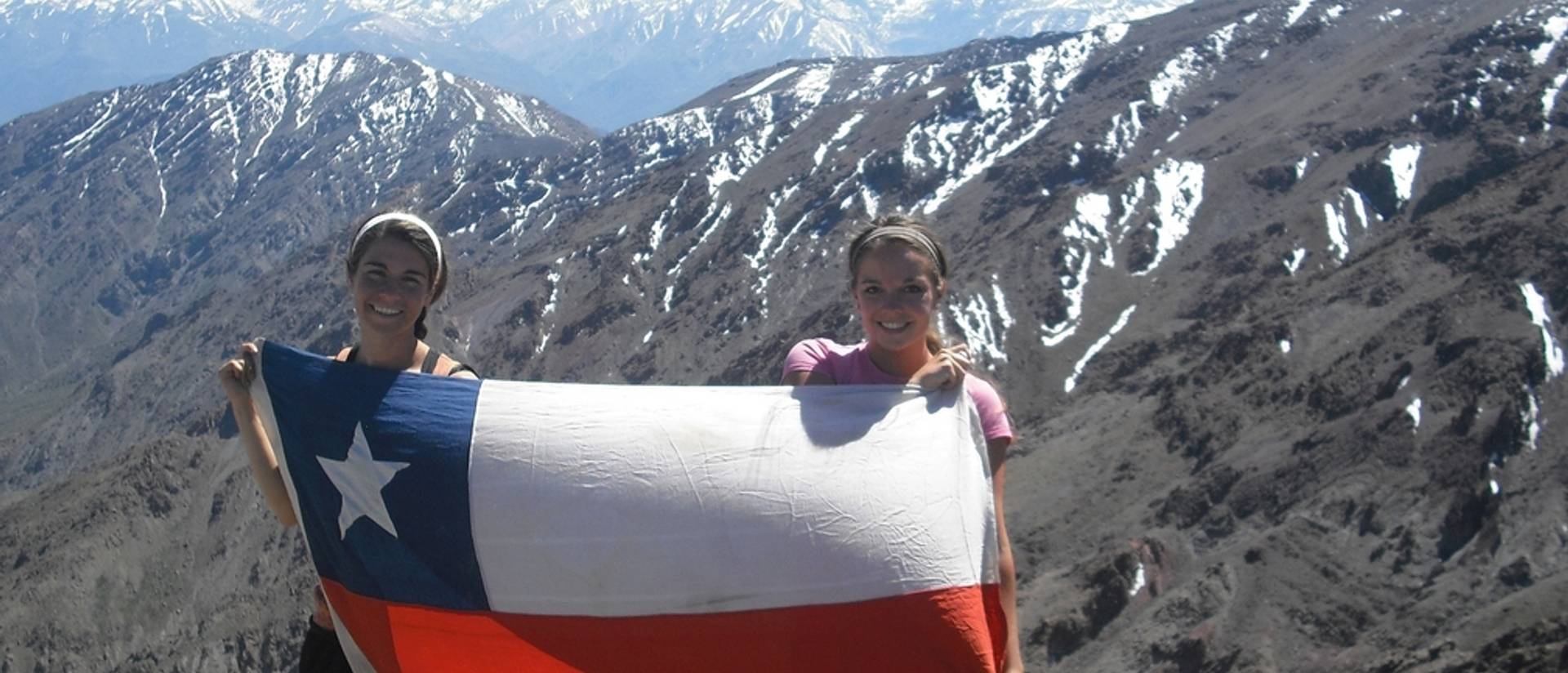 Two people smile while holding Chile's flag with mountains in the background.