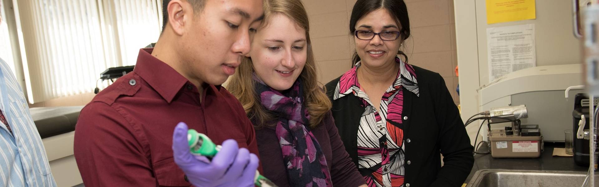 Three people in a chemistry lab look intently at the work being performed on a lab table.