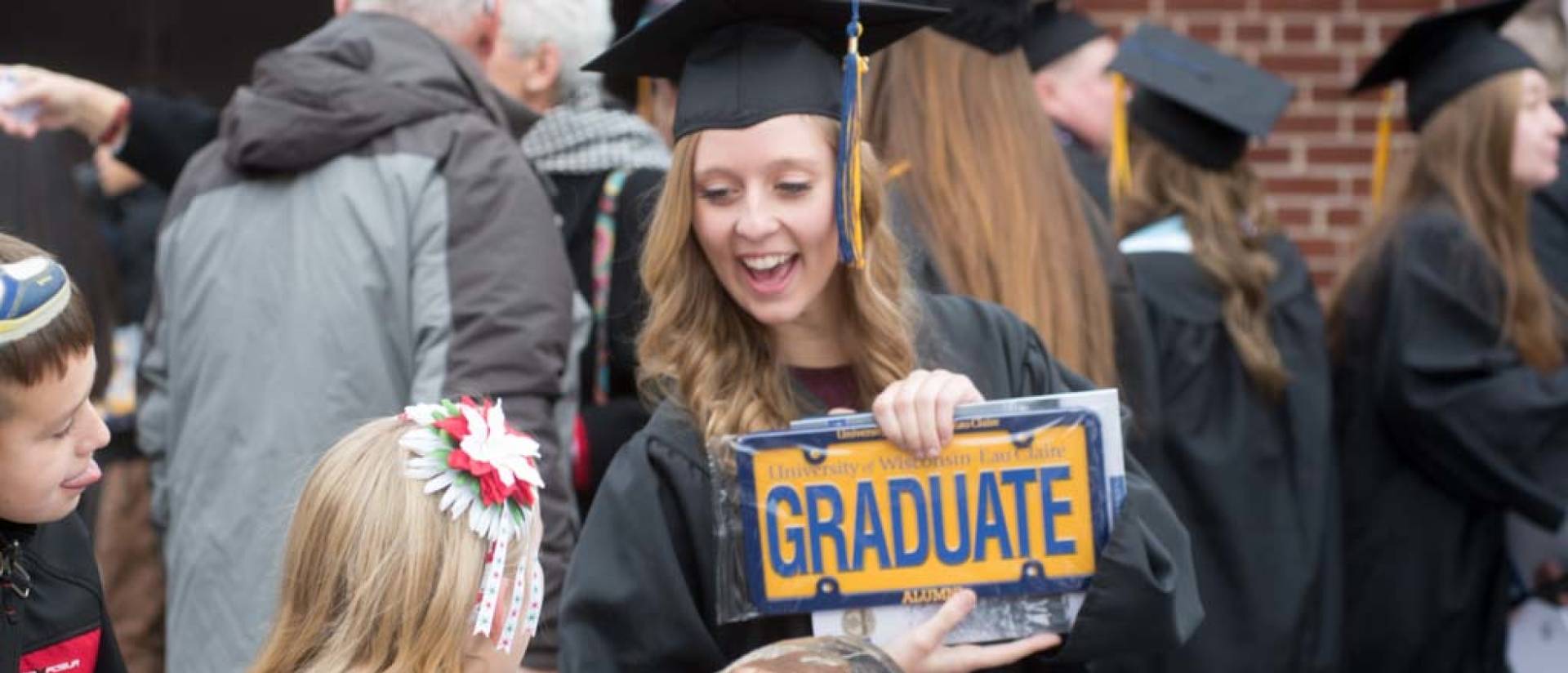 Student outside at commencement holding license plate that says “graduate”