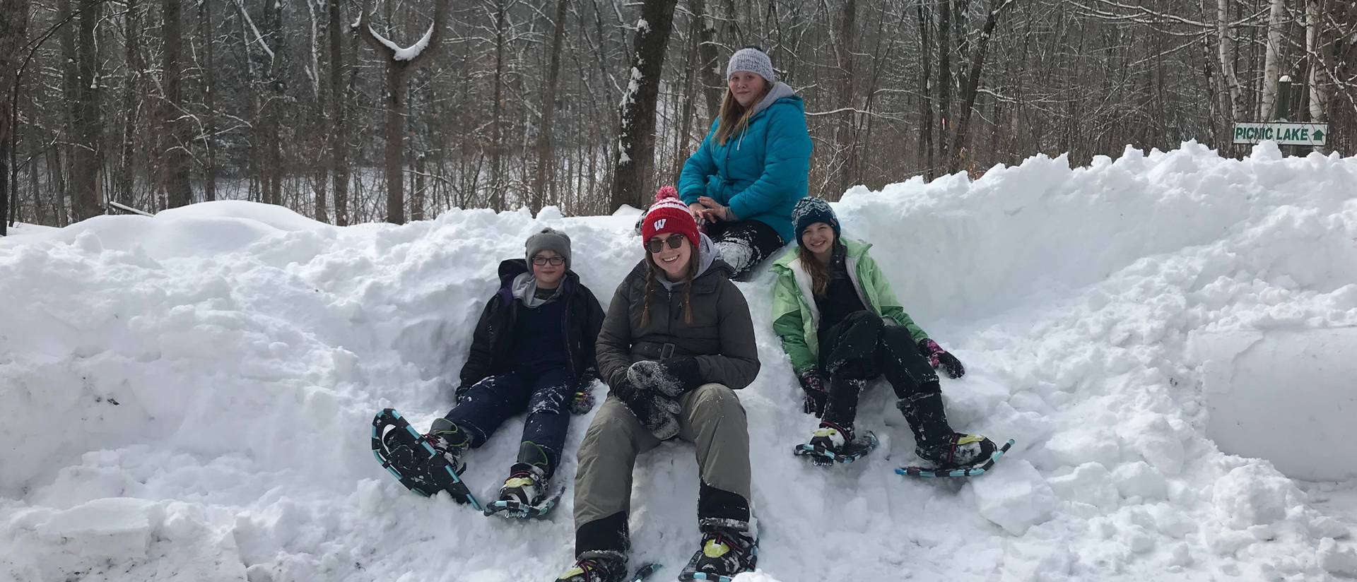 Four people wearing snowshoes sit on a snowbank and smile.